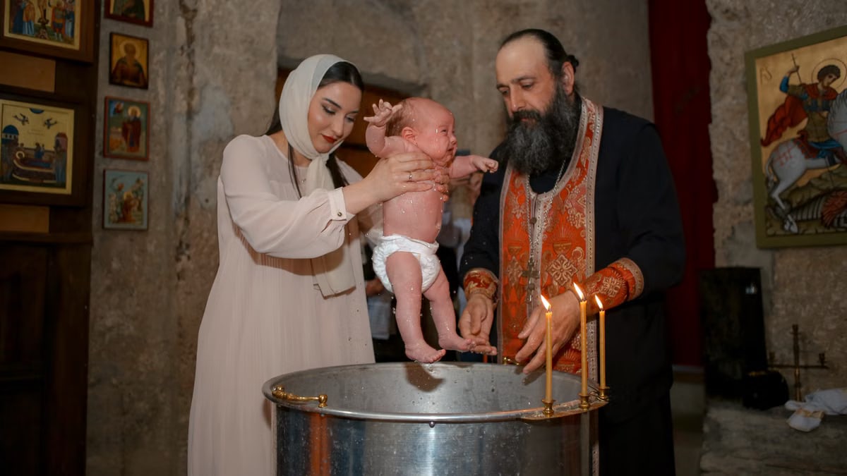 A baby being baptized by a priest and woman in a traditional church setting.